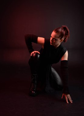 Siyah fütüristik deri pelerin giyen güzel kızıl saçlı kadın modelin tam boy portresi. Karanlık stüdyo arka planında oturan karamsar ışıklandırmalı..