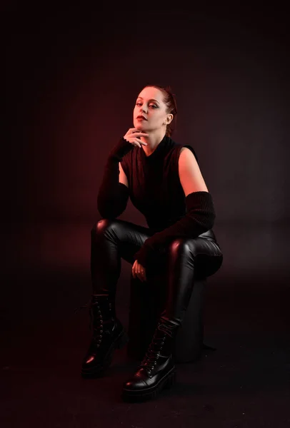 全长肖像漂亮红头发女模特身穿黑色未来科幻皮衣 坐在黑暗的摄影棚背景上 阴影环绕着情绪的灯光 — 图库照片