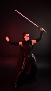 Siyah fütüristik deri pelerin giyen güzel kızıl saçlı kadın modelin tam boy portresi. Karanlık stüdyo arka planında, karamsar ışıklandırmalı ışın kılıcı tutan duruş..