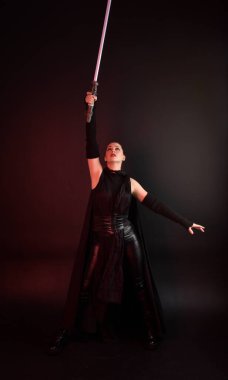 Siyah fütüristik deri pelerin giyen güzel kızıl saçlı kadın modelin tam boy portresi. Karanlık stüdyo arka planında, karamsar ışıklandırmalı ışın kılıcı tutan duruş..