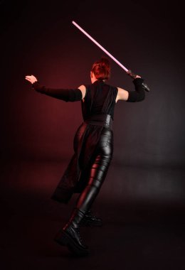 Siyah fütüristik deri pelerin giyen güzel kızıl saçlı kadın modelin tam boy portresi. Karanlık stüdyo arka planında, karamsar ışıklandırmalı ışın kılıcı tutan duruş..
