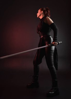 Siyah fütüristik deri pelerin giyen güzel kızıl saçlı kadın modelin tam boy portresi. Karanlık stüdyo arka planında, karamsar ışıklandırmalı ışın kılıcı tutan duruş..