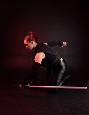 Siyah fütüristik deri pelerin giyen güzel kızıl saçlı kadın modelin tam boy portresi. Karanlık stüdyo arka planında, karamsar ışıklandırmalı ışın kılıcı tutan duruş..