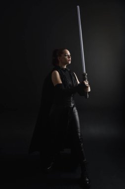 Siyah fütüristik deri pelerin giyen güzel kızıl saçlı kadın modelin tam boy portresi. Karanlık stüdyo arka planında, karamsar ışıklandırmalı ışın kılıcı tutan duruş..