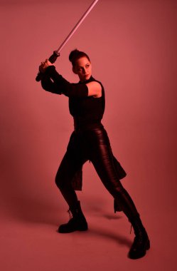 Siyah fütüristik deri pelerin giyen güzel kızıl saçlı kadın modelin tam boy portresi. Kırmızı stüdyo arka planında renkli ışıklandırmalı ışın kılıcı tutan duruş.