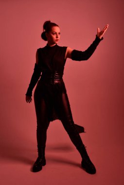 Siyah fütüristik deri pelerin giyen güzel kızıl saçlı kadın modelin tam boy portresi. Kırmızı stüdyo arka planında renkli ışıklandırmalı ışın kılıcı tutan duruş.