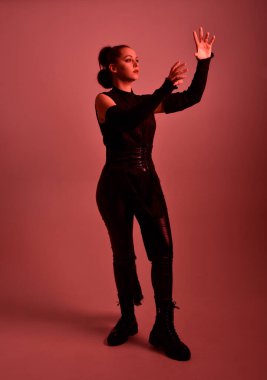 Siyah fütüristik deri pelerin giyen güzel kızıl saçlı kadın modelin tam boy portresi. Kırmızı stüdyo arka planında renkli ışıklandırmalı ışın kılıcı tutan duruş.