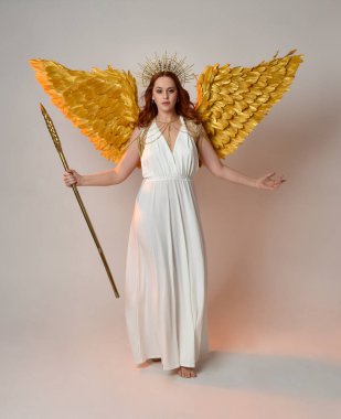 Kızıl saçlı güzel bir kadının portresi uzun, fantezi bir toga elbiseyle altın bir halo taç takıyor, beyaz bir stüdyonun arka planında izole edilmiş bir şekilde duruyor..