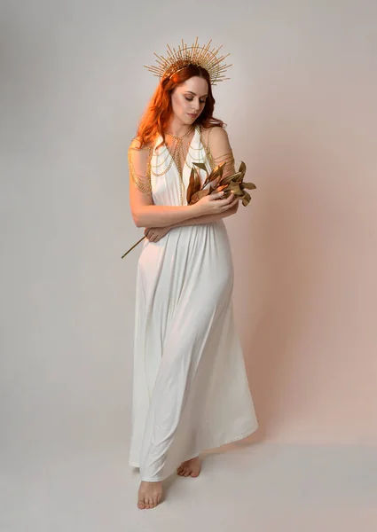 portrait of beautiful red head woman wearing long flowing fantasy toga gown with golden halo crown jewellery,  posed on isolated on a white studio background.