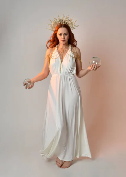 stock image portrait of beautiful red head woman wearing long flowing fantasy toga gown with golden halo crown jewellery,  posed on isolated on a white studio background.