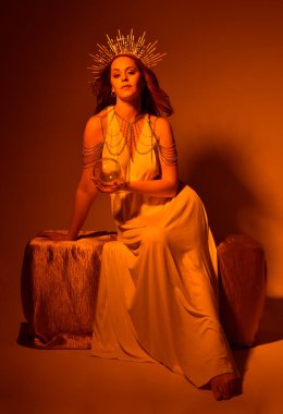 portrait of beautiful red head woman wearing long flowing fantasy toga gown with angelic  golden halo crown on studio background