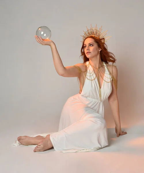 portrait of beautiful red head woman wearing long flowing fantasy toga gown with angelic  golden halo crown on studio background