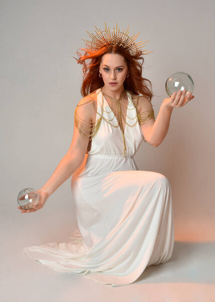 portrait of beautiful red head woman wearing long flowing fantasy toga gown with angelic  golden halo crown on studio background