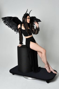 full length portrait of beautiful asian model with dark hair, wearing black gothic skirt costume, angel feather wings with horned headdress. Sitting pose  isolated on studio background.