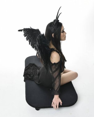 full length portrait of beautiful asian model with dark hair, wearing black gothic skirt costume, angel feather wings with horned headdress. kneeling  pose  isolated on studio background.