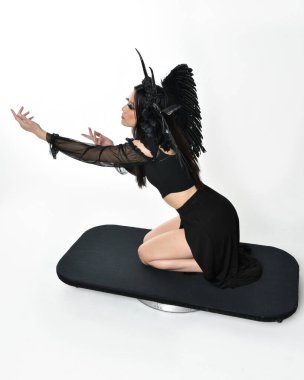 full length portrait of beautiful asian model with dark hair, wearing black gothic skirt costume, angel feather wings with horned headdress. kneeling  pose  isolated on studio background.