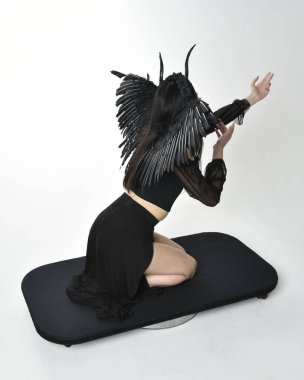 full length portrait of beautiful asian model with dark hair, wearing black gothic skirt costume, angel feather wings with horned headdress. kneeling  pose  isolated on studio background.