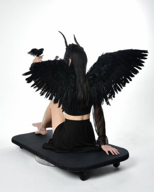 full length portrait of beautiful asian model with dark hair, wearing black gothic skirt costume, angel feather wings with horned headdress. kneeling  pose  isolated on studio background.