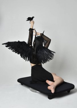full length portrait of beautiful asian model with dark hair, wearing black gothic skirt costume, angel feather wings with horned headdress. kneeling  pose  isolated on studio background.