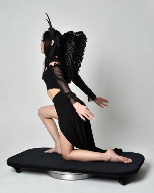 full length portrait of beautiful asian model with dark hair, wearing black gothic skirt costume, angel feather wings with horned headdress. kneeling  pose  isolated on studio background.