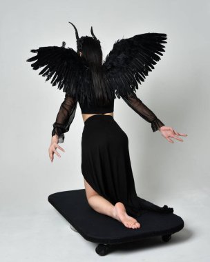 full length portrait of beautiful asian model with dark hair, wearing black gothic skirt costume, angel feather wings with horned headdress. kneeling  pose  isolated on studio background.