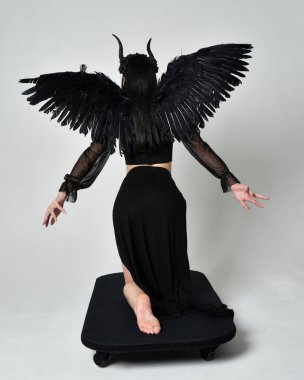 full length portrait of beautiful asian model with dark hair, wearing black gothic skirt costume, angel feather wings with horned headdress. kneeling  pose  isolated on studio background.