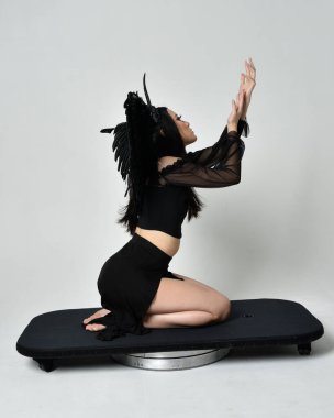 full length portrait of beautiful asian model with dark hair, wearing black gothic skirt costume, angel feather wings with horned headdress. kneeling  pose  isolated on studio background.