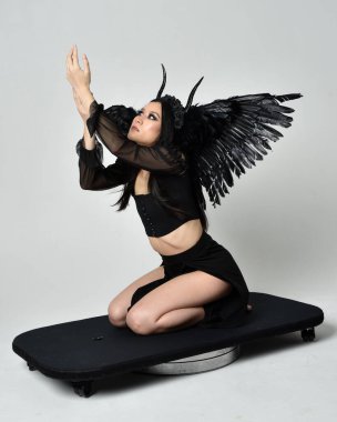full length portrait of beautiful asian model with dark hair, wearing black gothic skirt costume, angel feather wings with horned headdress. kneeling  pose  isolated on studio background.