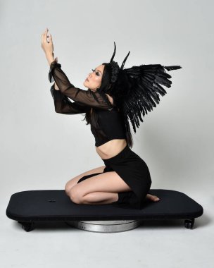 full length portrait of beautiful asian model with dark hair, wearing black gothic skirt costume, angel feather wings with horned headdress. kneeling  pose  isolated on studio background.