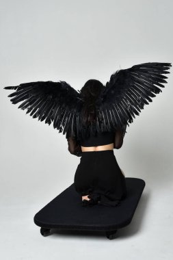 full length portrait of beautiful asian model with dark hair, wearing black gothic skirt costume, angel feather wings with horned headdress. kneeling  pose  isolated on studio background.