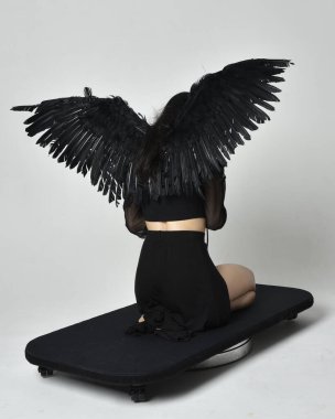 full length portrait of beautiful asian model with dark hair, wearing black gothic skirt costume, angel feather wings with horned headdress. kneeling  pose  isolated on studio background.