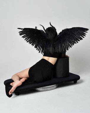 full length portrait of beautiful asian model with dark hair, wearing black gothic skirt costume, angel feather wings with horned headdress. kneeling  pose  isolated on studio background.