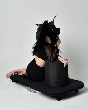 full length portrait of beautiful asian model with dark hair, wearing black gothic skirt costume, angel feather wings with horned headdress. kneeling  pose  isolated on studio background.