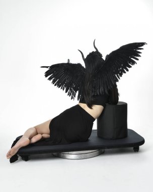 full length portrait of beautiful asian model with dark hair, wearing black gothic skirt costume, angel feather wings with horned headdress. kneeling  pose  isolated on studio background.