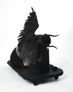 full length portrait of beautiful asian model with dark hair, wearing black gothic skirt costume, angel feather wings with horned headdress. kneeling  pose  isolated on studio background.