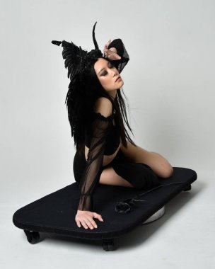 full length portrait of beautiful asian model with dark hair, wearing black gothic skirt costume, angel feather wings with horned headdress. kneeling  pose  isolated on studio background.