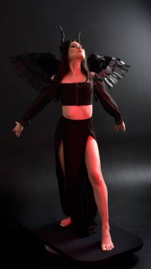 portrait of beautiful asian model with dark hair, wearing black gothic skirt costume, angel feather wings with horned headdress. Posing with gestural hands  on dark silhouette  studio background.