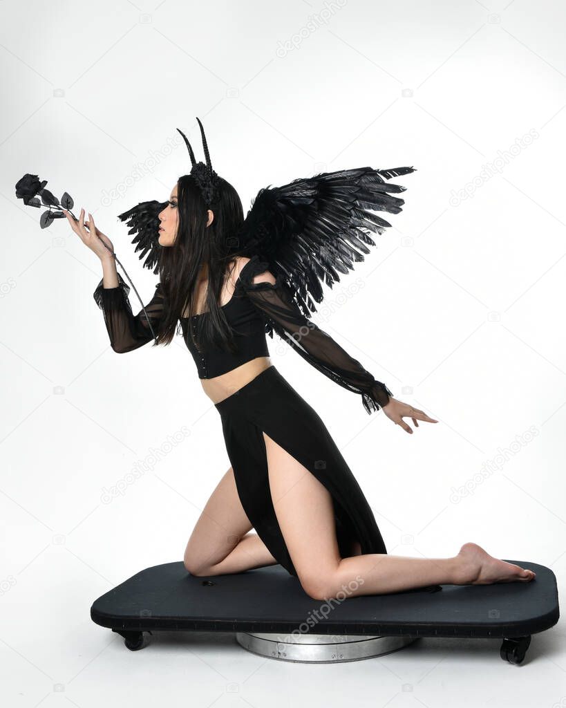 full length portrait of beautiful asian model with dark hair, wearing black gothic skirt costume, angel feather wings with horned headdress. kneeling  pose  isolated on studio background.