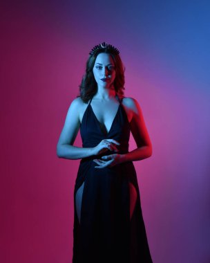 Zarif siyah elbise ve taç giyen güzel bir kadın modelin yakın plan portresi, fantezi dolu kol hareketleriyle bir stüdyo arka planına poz vermek, çok renkli yaratıcı ışıklandırma..