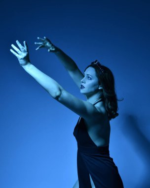 Zarif siyah elbise ve taç giyen güzel bir kadın modelin yakın plan portresi, fantezi dolu kol hareketleriyle bir stüdyo arka planına poz vermek, çok renkli yaratıcı ışıklandırma..