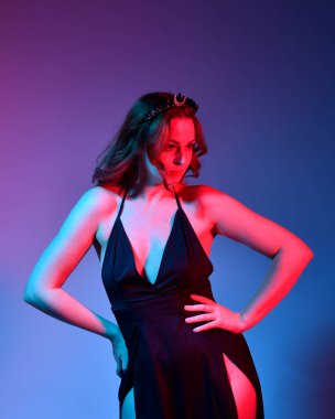 Zarif siyah elbise ve taç giyen güzel bir kadın modelin yakın plan portresi, fantezi dolu kol hareketleriyle bir stüdyo arka planına poz vermek, çok renkli yaratıcı ışıklandırma..