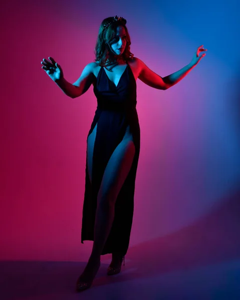 Close up portrait of beautiful woman model wearing elegant black dress and crown, posing against a studio background with fantasy inspired arm gestures, multi coloured creative lighting.