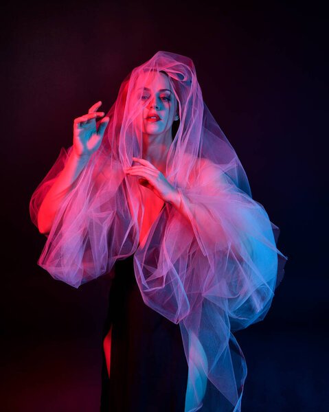 Крупный план портрета красивой женщины-модели в элегантном черном платье и марлевой саван ткани, позируя на фоне студии с фантазией вдохновенных жестов руки, разноцветного творческого освещения.