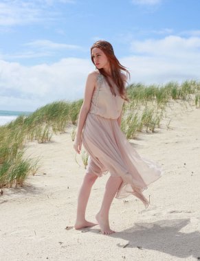 Uzun saçlı, rüzgarda süzülen basit bir elbise giyen güzel bir genç kadının tam boy portresi. Zarif bir duruş. Okyanus sahili arka planında altın günbatımı ışığı.