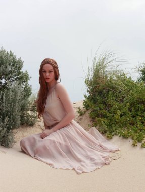 Uzun saçlı, rüzgarda süzülen basit bir elbise giyen güzel bir genç kadının tam boy portresi. Zarif bir duruş. Okyanus sahili arka planında altın günbatımı ışığı.