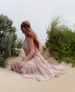 Uzun saçlı, rüzgarda süzülen basit bir elbise giyen güzel bir genç kadının tam boy portresi. Zarif bir duruş. Okyanus sahili arka planında altın günbatımı ışığı.