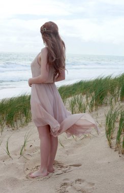 Uzun saçlı, rüzgarda süzülen basit bir elbise giyen güzel bir genç kadının tam boy portresi. Zarif bir duruş. Okyanus sahili arka planında altın günbatımı ışığı.