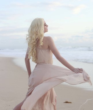 Uzun saçlı, rüzgarda süzülen basit bir elbise giyen güzel bir genç kadının tam boy portresi. Zarif bir duruş. Okyanus sahili arka planında altın günbatımı ışığı.