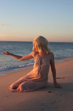 Uzun saçlı, rüzgarda süzülen basit bir elbise giyen güzel bir genç kadının tam boy portresi. Zarif bir duruş. Okyanus sahili arka planında altın günbatımı ışığı.