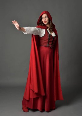 Uzun etekli ve dalgalı kapüşonlu pelerinli kırmızı ortaçağ fantezi kostümü giyen güzel esmer kadının tam boy portresi. El hareketleriyle poz veriyor, gri stüdyo arka planında izole edilmiş..